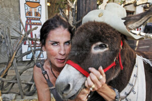 Animals Birds Insects Eagles Geese Horse Plants, A smiling woman with dark hair embraces an animal, a donkey wearing a light-colored hat. She holds the donkey's face close to hers. The animal is wearing a red halter. Behind them, a rustic wooden fence and a Native American artwork provide a backdrop to the scene. pictures images stock photography getty shutterstock